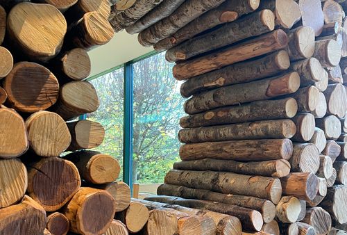 Foto mit aufgeschichteten Rundhölzern, die einen runden Durchblick freilassen in einen angrenzenden Raum. Dort sieht man ein Fenster und dahinter Bäume.