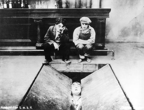 Charlie Chaplin und eine Frau sitzen nebeneinander vor einer Theke auf dem Boden. Vor ihnen tut sich eine große, sich nach unten öffnende Doppel-Falltür auf, aus der ein Mann schaut.