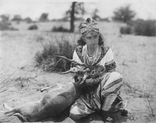 Ein schwarz-weißes Foto. Ein Mann in traditioneller indischer Tracht sitzt im Freien auf sandigem Boden. Er hält ein möglicherweise totes Tier: eine Art Gämse.